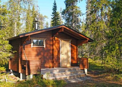 Punaruskeaseinäinen, hirsinen saunarakennus kesämaisemassa.