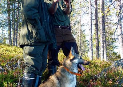 Kaksi miestä kameroineen ja koira rinteessä.
